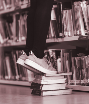 Person steht auf einem Bücherstapel, um an das obere Bücherregal zu kommen